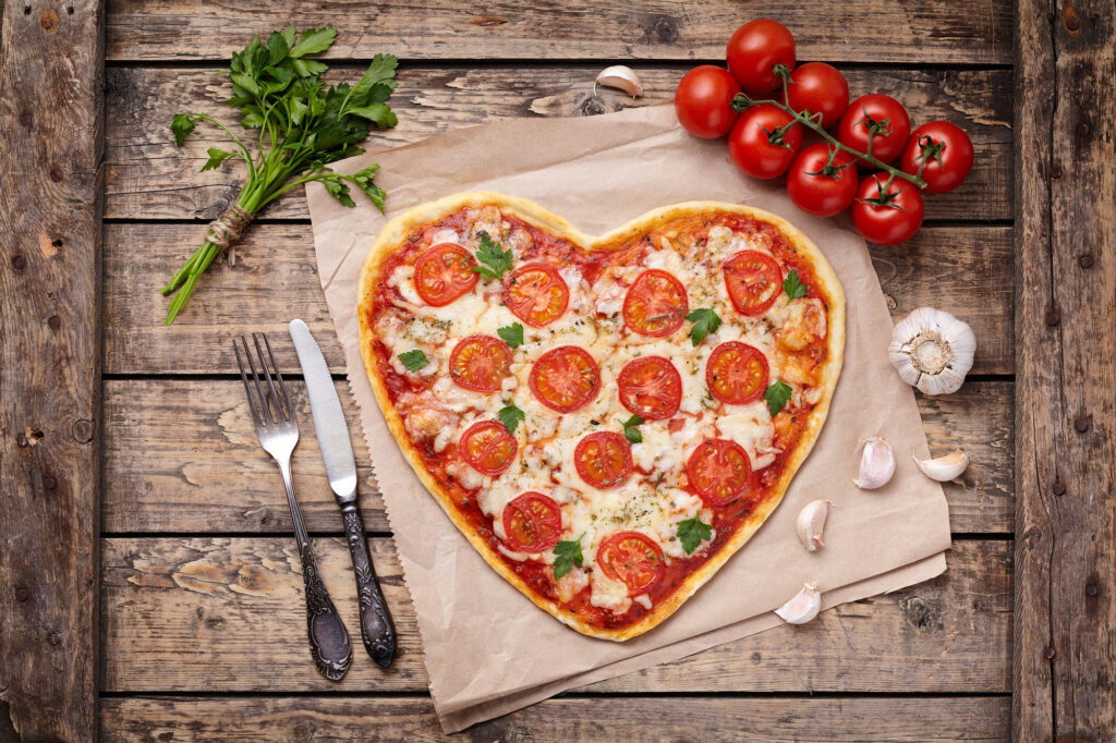 Heart shaped pizza margherita love concept for Valentines Day with mozzarella, tomatoes, parsley and garlic on vintage wooden table background. Rustic style and top view.